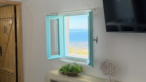 Ein Zimmer mit Holztür, einem Fenster mit Meerblick, einem an der Wand befestigten Flachbildfernseher und einer kleinen Pflanze auf einem Beistelltisch.