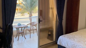 Una habitación de hotel con cama, puerta corrediza al balcón, ventilador, mesa con sillas y vistas a edificios y palmeras.
