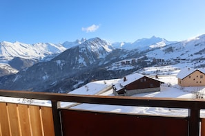Une vue sur les montagnes, avec des sommets enneigés, un balcon en bois et plusieurs bâtiments dans la vallée.