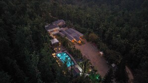An aerial view of a resort with multiple buildings, a swimming pool, and a large parking area surrounded by dense forest. - KAVA RETREAT (Chikkamagaluru)