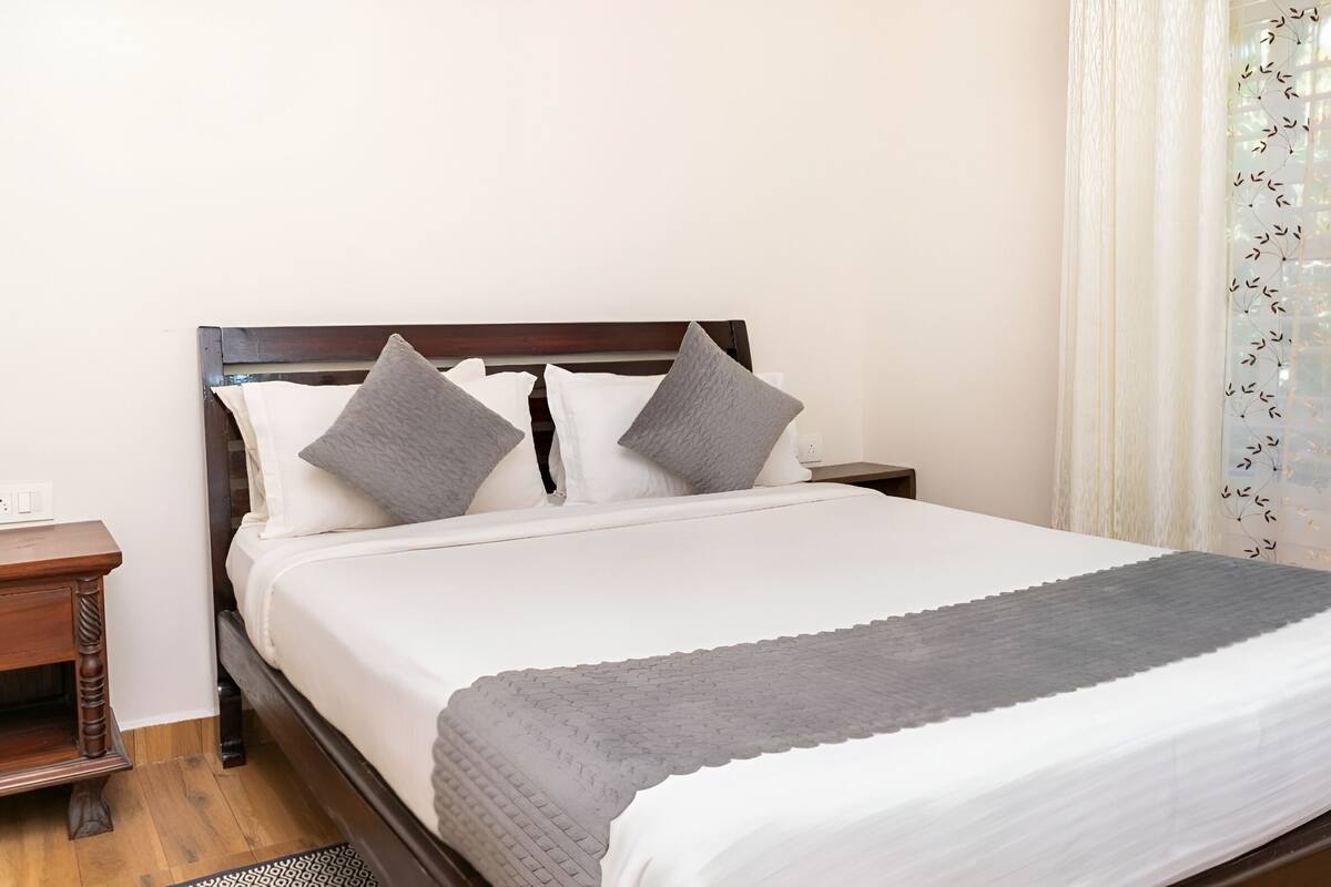 A neatly made bed with a grey blanket, two pillows, a wooden headboard, a bedside table with a lamp, and a window with a curtain.
