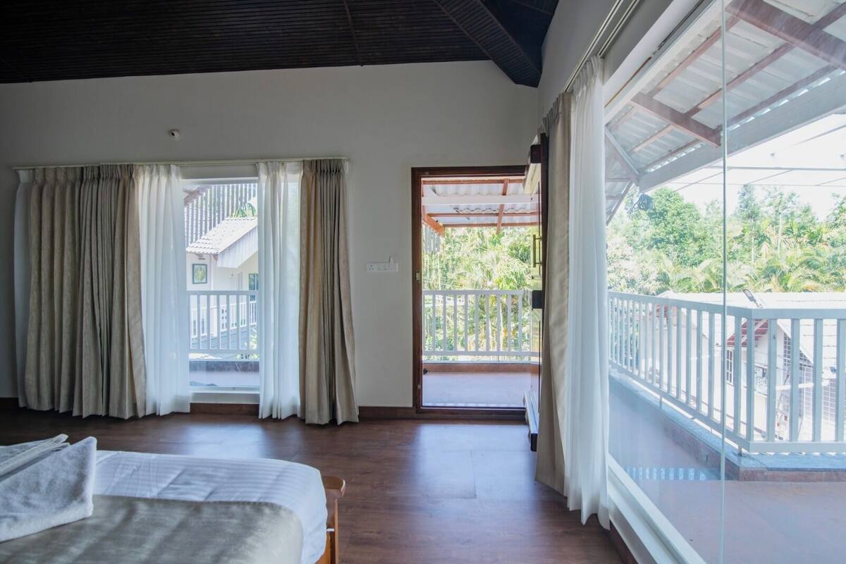 A room with a bed, curtains, a balcony with a railing, and a view of greenery outside.