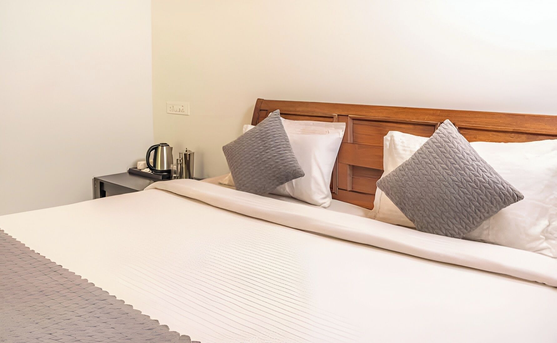 Una cama bien hecha con cabecera de madera, una mesita de noche con hervidor y un enchufe en la pared.