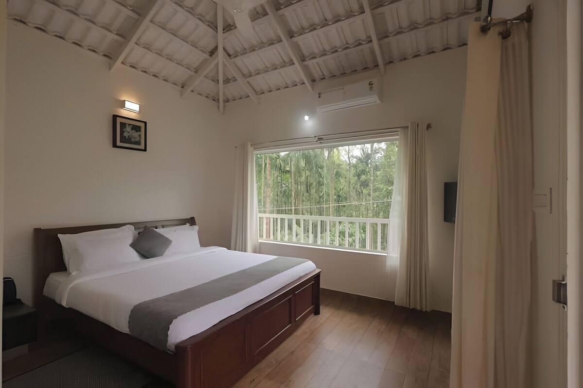 A bedroom with a bed, a window with a view of greenery, a ceiling fan, and a light fixture.