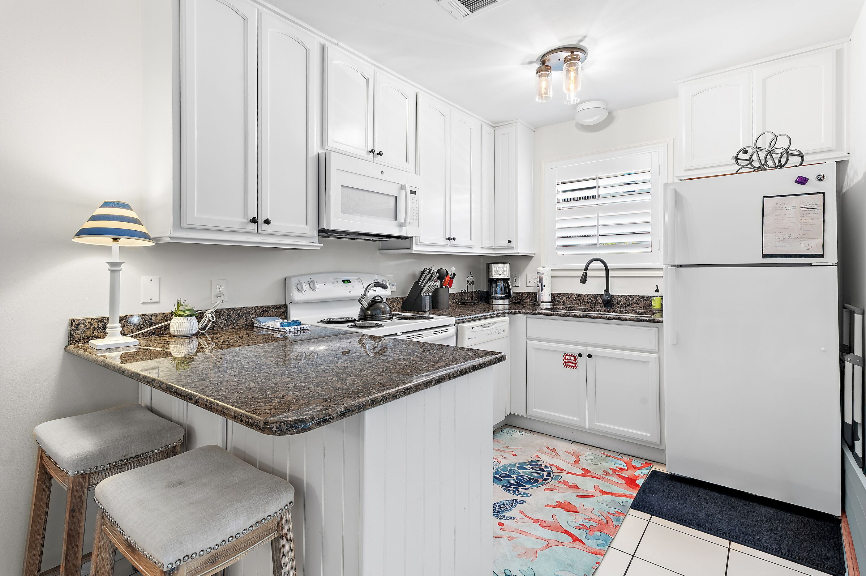 A kitchen with white cabinets, a granite countertop, a stove, a sink, and a refrigerator. There are two stools and a lamp.