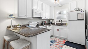 Una cocina con armarios blancos, encimera de granito, estufa, fregadero y refrigerador. Hay dos taburetes y una lámpara.