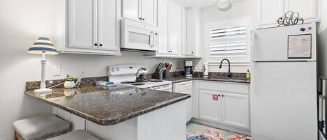 A kitchen with white cabinets, a granite countertop, a stove, a sink, and a refrigerator. There are two stools and a lamp.