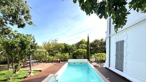 En rektangulär swimmingpool omgiven av ett trädäck, en pergola och en vit byggnad med fönsterluckor i bakgrunden.