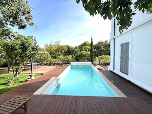 Una piscina rectangular con una terraza de madera alrededor, una pérgola y un edificio blanco con persianas al fondo.