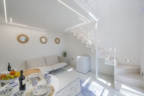 A modern living room with a white sofa, a dining table set with plates and glasses, a refrigerator, and a staircase. - Casa Lucente - terrace sea view (Montepertuso)