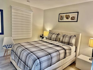 A bedroom with a bed, a chair, a bedside table, a lamp, a wall-mounted picture, and a window with blinds. - Airport Urban Oasis 4BR Family Retreat Home (Dieppe)