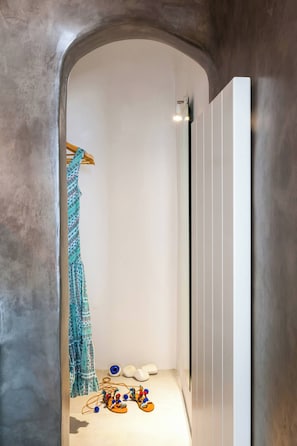 Un couloir étroit avec une arche en béton, une robe à motifs suspendue et une paire de chaussures colorées posées sur le sol.