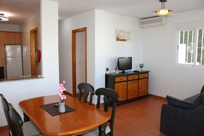 Ein Essbereich mit Holztisch, Stühlen, einer kleinen Vase mit Blumen, einem Fernseher auf einem Schrank und einem Fenster mit Gitterstäben.