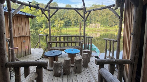 Una terraza de madera con una estructura rústica de madera, un pequeño edificio de madera con el letrero 'Cottage' y vistas a un lago y un bosque.