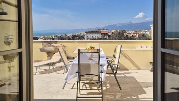 Ein Balkon mit Tisch und Stühlen, Blick auf eine Küstenstadt und ein Gebirge im Hintergrund.