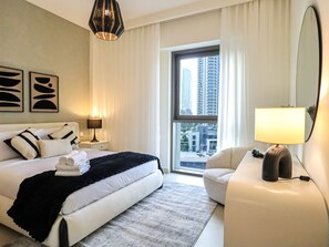 A hotel room with a bed, a chair, a desk, a lamp, a window with a city view, and abstract wall art. - 2BR  Apt in Dubai Creek Grove 3 (Dubai)
