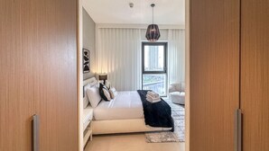 A modern bedroom with a large bed, a view of a cityscape through a window, and a hanging light fixture. - 2BR  Apt in Dubai Creek Grove 3 (Dubai)