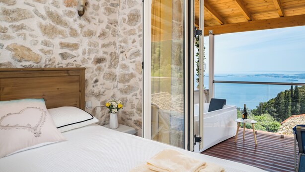 A bedroom with a stone wall, a wooden headboard, a bed with white bedding, a bedside table with a vase of flowers, and a balcony with a view of the sea. - Art House with Seaview by Konnect, Spartylas (Corfu)