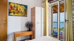 A bedroom with a bed, a wooden bedside table, a fan, a wardrobe, a painting, and a sliding glass door leading to a balcony with a view of the sea. - Art House with Seaview by Konnect, Spartylas (Corfu)