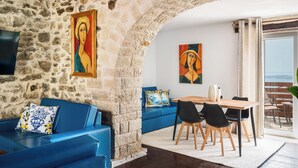A stone-walled room with a blue sofa, a wooden dining table with chairs, and artwork on the walls. - Art House with Seaview by Konnect, Spartylas (Corfu)