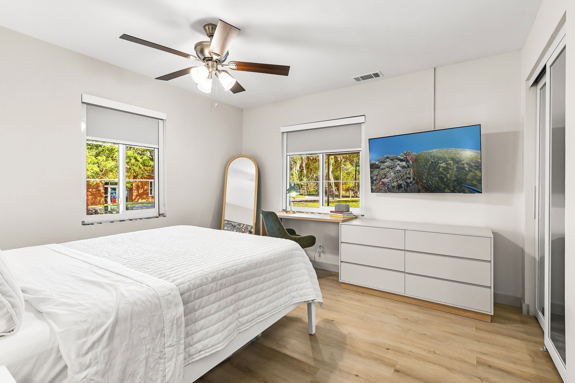 A bedroom with a bed, a desk, a chair, a TV, and two windows with views of trees.
