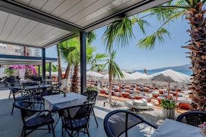 An outdoor seating area with tables and chairs, palm trees, and umbrellas overlooking a beach.