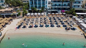 A beach with sun loungers and umbrellas, a pool, and a building in the background. - Vila Era Beach 2 (Sarande)