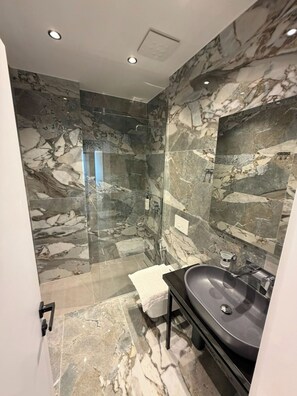 A modern bathroom with a glass-enclosed shower, marble walls, and a black sink. - Vila Era Beach 2 (Sarande)