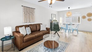 A living room with a brown leather sofa, a glass coffee table, a ceiling fan, a macrame wall hanging, and a dining area with blue chairs. - Park the car and enjoy Mount Dora! <br>Stay just a 1/2 Mile from Downtown Mount Dora and Lake Dora in our beautifully restored 1950s duplex. <br>Walk to shops & dining! Features fast WiFi, workspace, patio, free parking, and boat parking! (Mount Dora)