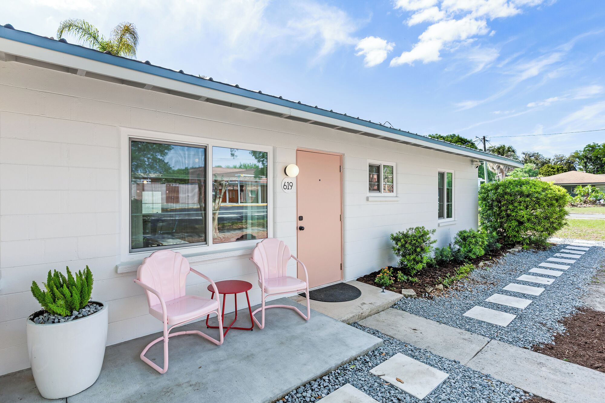 A single-story building with a white exterior, a pink door, and a number '619' on the wall. There are two pink chairs outside, a potted plant, and a gravel pathway leading to the entrance.