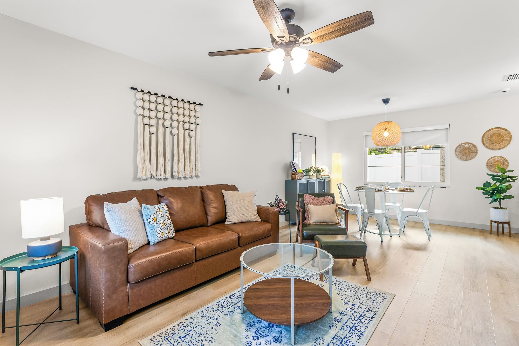 A modern living room with a brown leather sofa, a glass coffee table, a ceiling fan, and a dining area with a table and chairs.