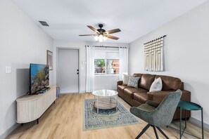 A living room with a brown leather sofa, a glass coffee table, a patterned rug, a ceiling fan, and a wall-mounted macrame piece. - Park the car and enjoy Mount Dora! <br>Stay just a 1/2 Mile from Downtown Mount Dora and Lake Dora in our beautifully restored 1950s duplex. <br>Walk to shops & dining! Features fast WiFi, workspace, patio, free parking, and boat parking! (Mount Dora)