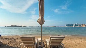 Una playa con dos sillas de playa y una sombrilla, un mar tranquilo y edificios a lo lejos.