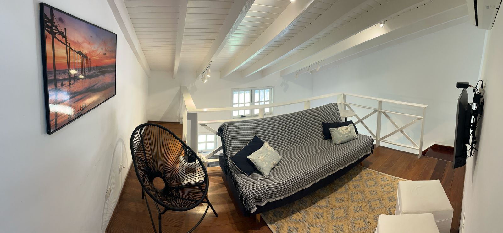 Una habitación estilo loft con techo alto, un sofá a rayas, una silla negra y un cuadro de un muelle.