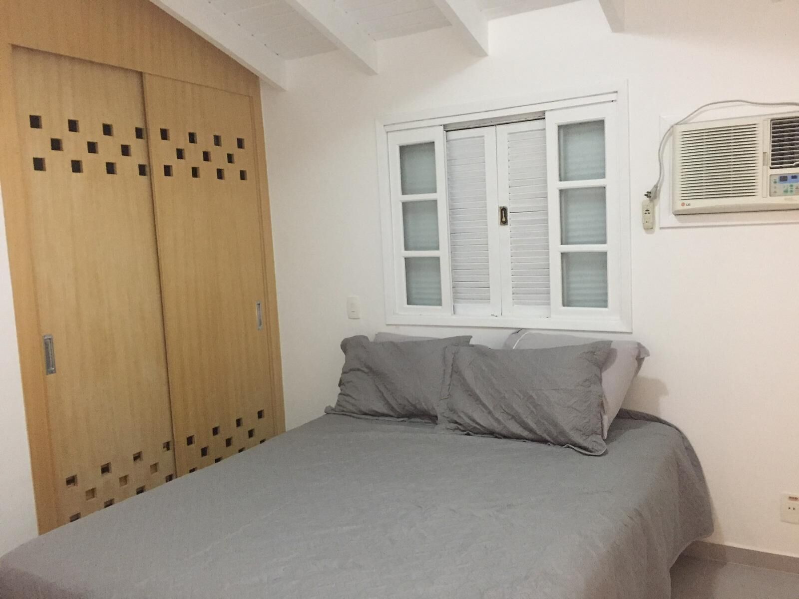 A bedroom with a bed, a window with shutters, a wall-mounted air conditioner, and a wooden wardrobe with a unique pattern.