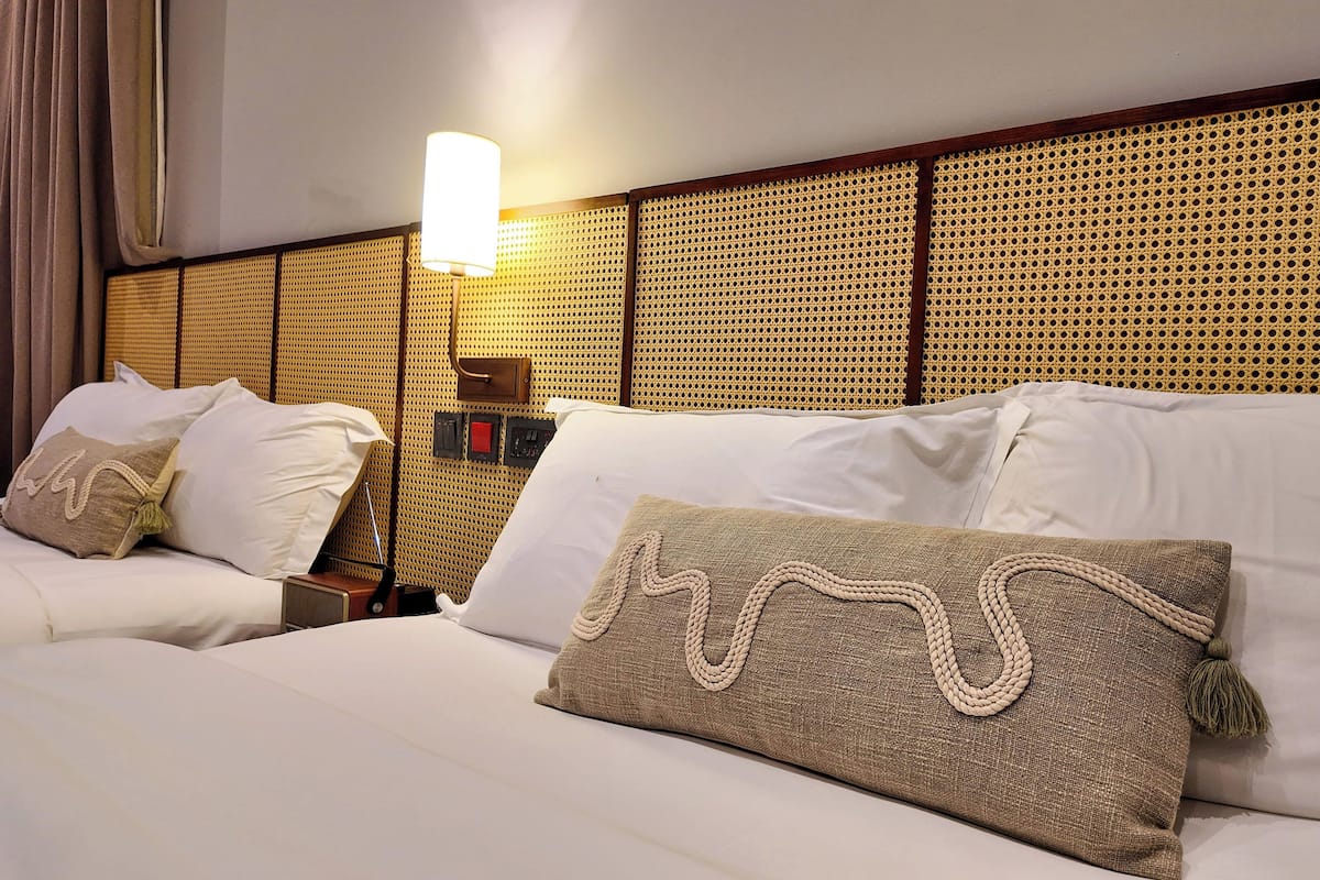 Ein Hotelzimmer mit zwei Betten, einem Kopfteil mit Webmuster, einem Nachttisch mit Lampe sowie einer an der Wand befestigten Schalterleiste.