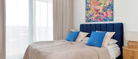 A bedroom with a bed, a large window with sheer curtains, a colorful abstract painting on the wall, and a wooden nightstand with a lamp.