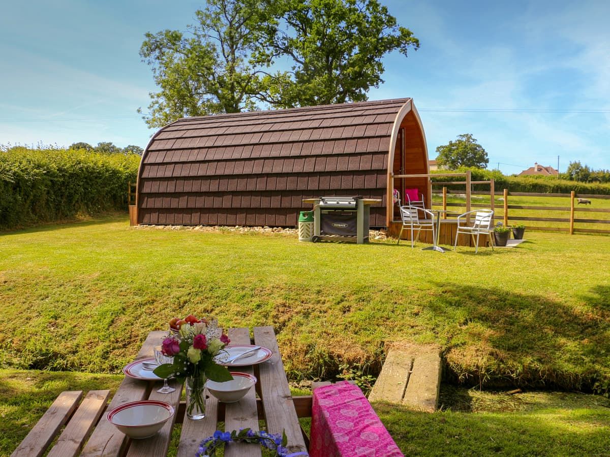 A wooden structure with a curved roof, a picnic table with plates and a vase of flowers, and a fenced area with chairs and a table.