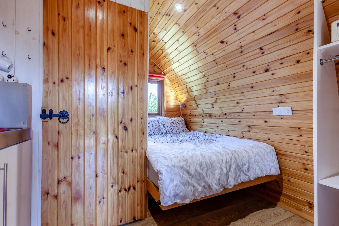 A bedroom with a sloped ceiling, wooden walls, a bed with a patterned bedspread, and a window with a view of greenery.