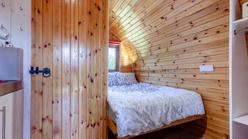 A bedroom with a sloped ceiling, wooden walls, a bed with a patterned bedspread, and a window with a view of greenery.