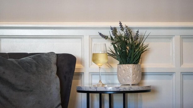 Une pièce avec un canapé gris, une petite table ronde ornée d’une plante et d’un vase, et un mur recouvert de lambris.