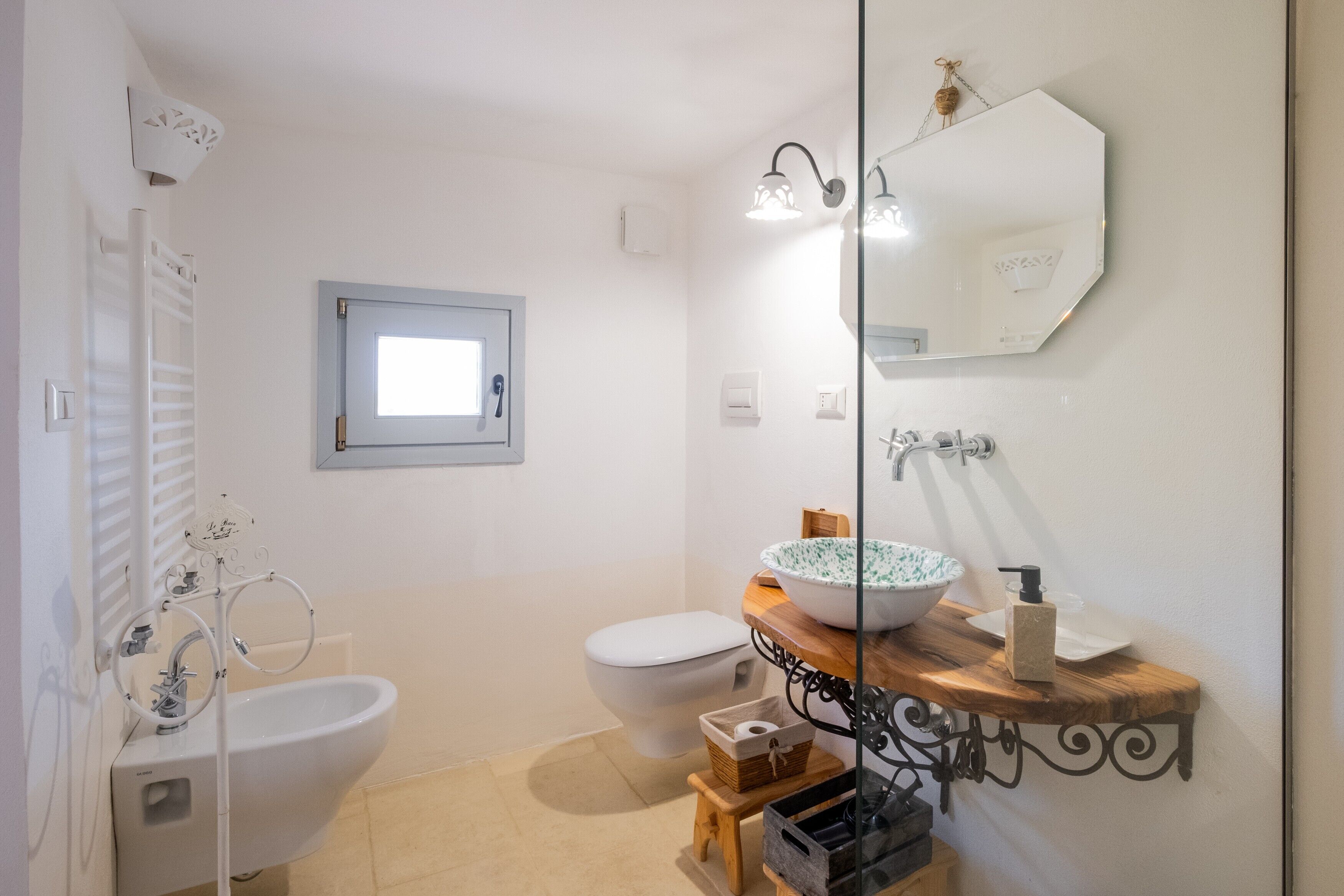A bathroom with a white sink, a white toilet, a bidet, a mirror, and a small window.