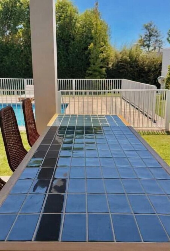 A patio with a tiled table and chairs, surrounded by a white fence and greenery.