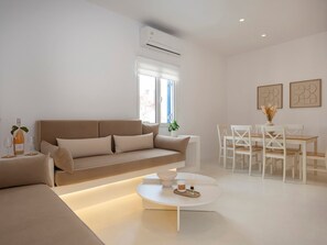 A modern living room with a sofa, a coffee table, a dining area with chairs, and a window with blinds. - Victoria s Relax House (Paros)