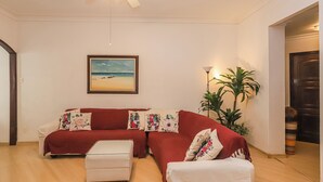 Un salon avec un canapé rouge, un pouf blanc, un ventilateur de plafond, un tableau représentant une plage, encadré, et une plante en pot.