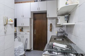 A compact kitchen with white cabinets, a wooden door, a stainless steel sink, and a glass-top stove. - Leme 2 quartos amplo 2 min praia (Rio de Janeiro)