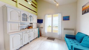 A room with a white cabinet, a teal sofa, a television, and a window with a view. - Spot de la Corniche - Oceanfront Apartment (Saint-Hilaire-de-Riez)
