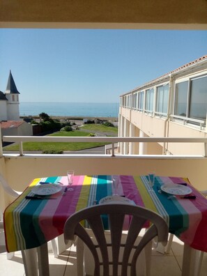 A balcony with a table set for two, overlooking a coastal view with a church and beach. - Spot de la Corniche - Oceanfront Apartment (Saint-Hilaire-de-Riez)