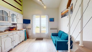Habitació estreta amb una cuineta, un sofà blau, una televisió i una vista per una porta.