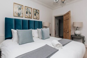 Una cama bien hecha con un cabecero azul, ropa de cama blanca y una toalla enrollada. Hay tres cuadros con motivos florales en la pared sobre la cama.